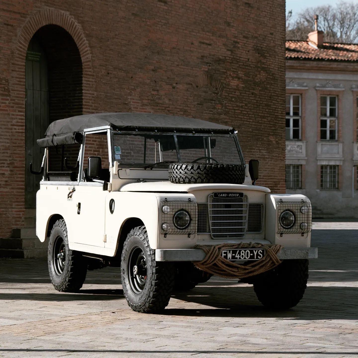 Un Land Rover vintage couleur crème est garé sur des pavés, avec un bâtiment en briques en arrière-plan.