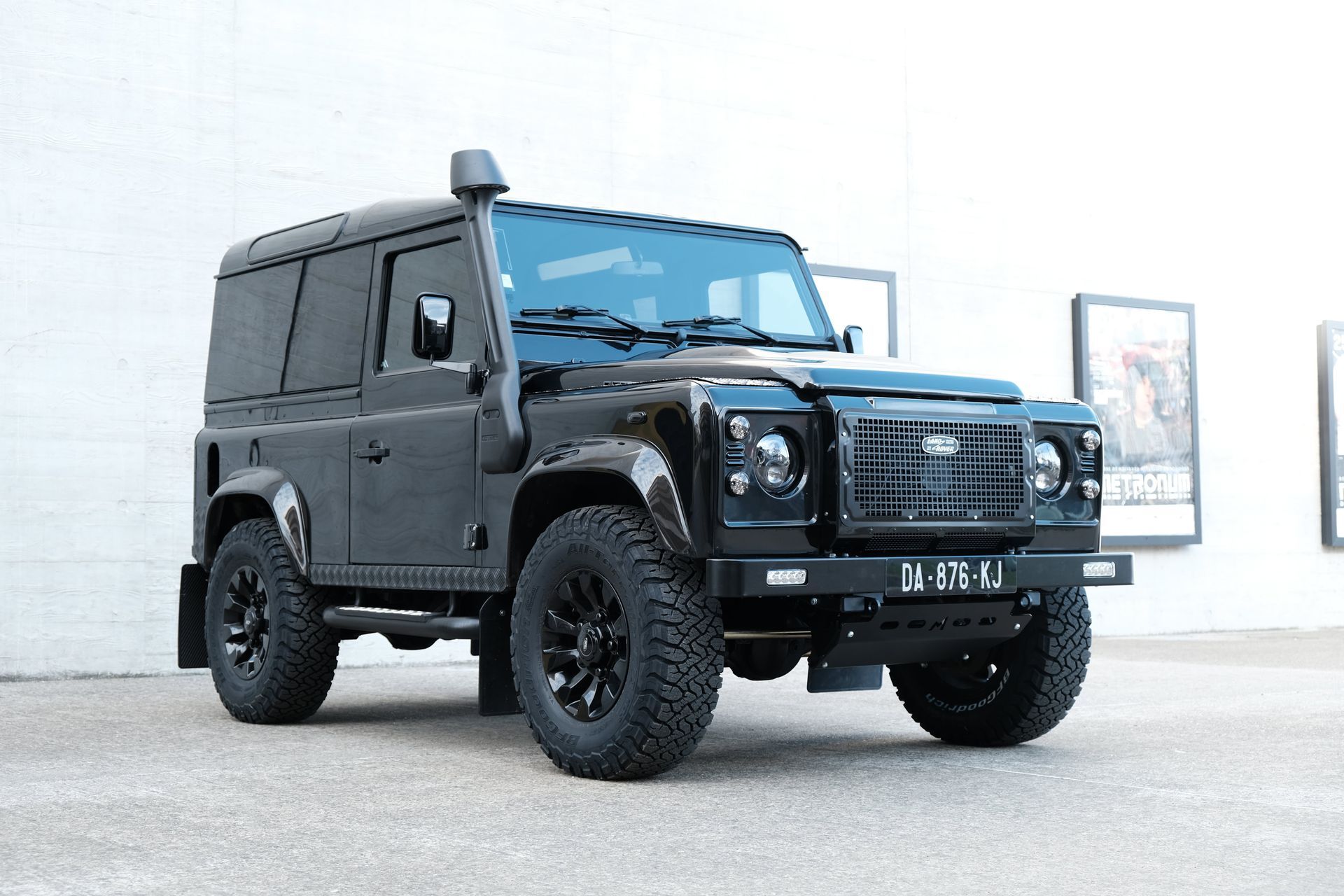 Un Land Rover Defender SUV noir, en angle, sur une surface pavée, contre un mur blanc.