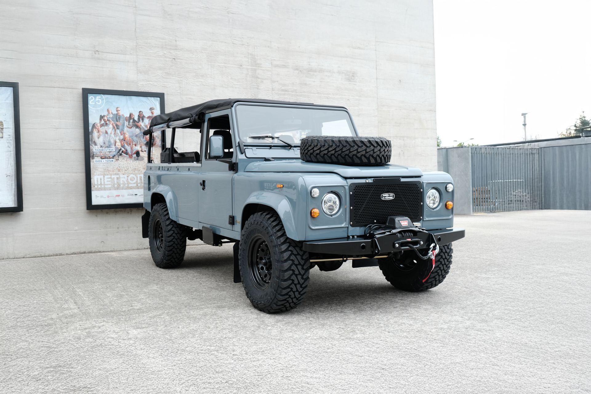 Land Rover Defender gris garé à l'extérieur, roue de secours sur le capot, treuil sur le pare-chocs avant.