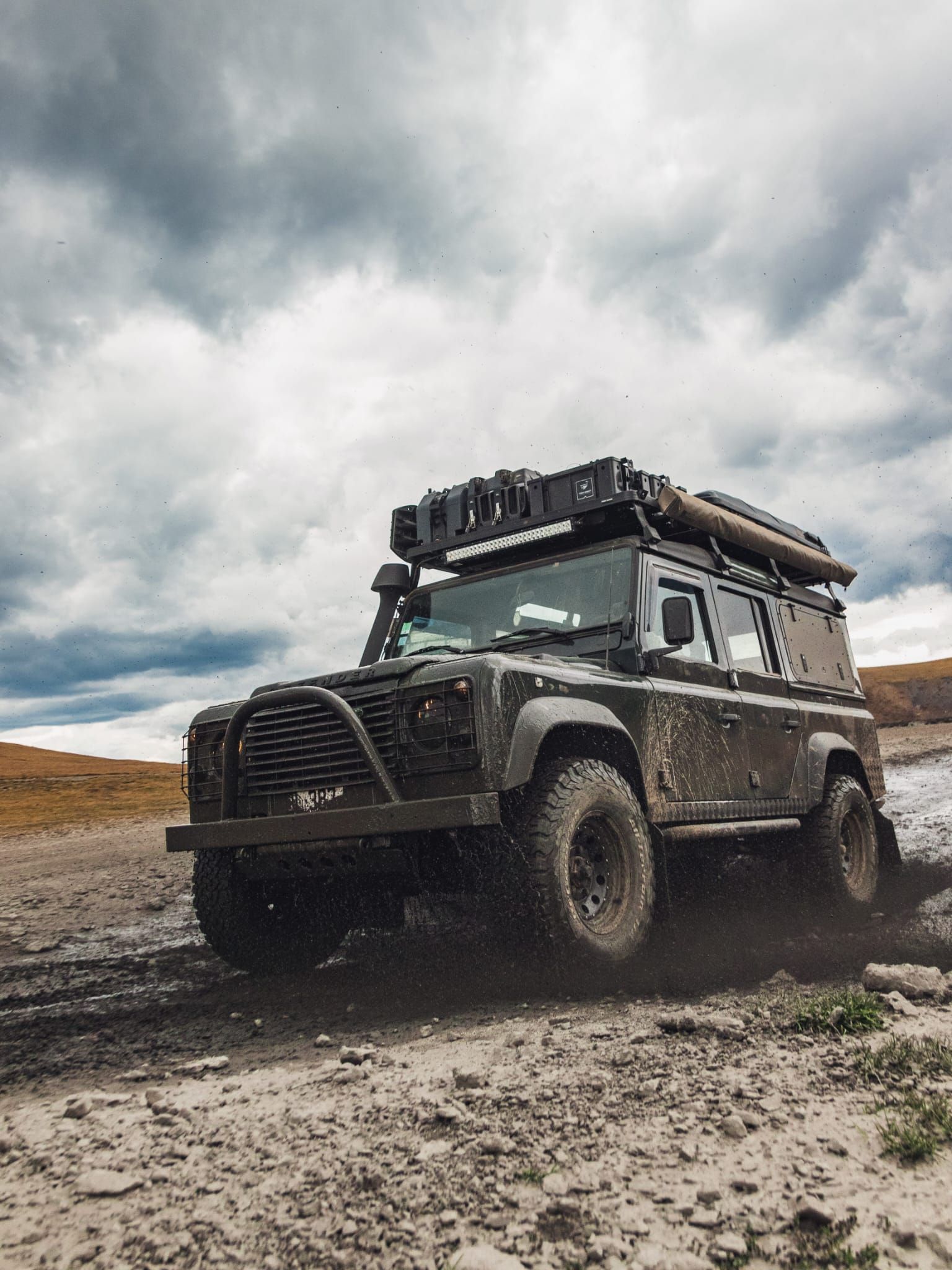 Un Land Rover Defender vert foncé roule sur un terrain boueux sous un ciel nuageux.