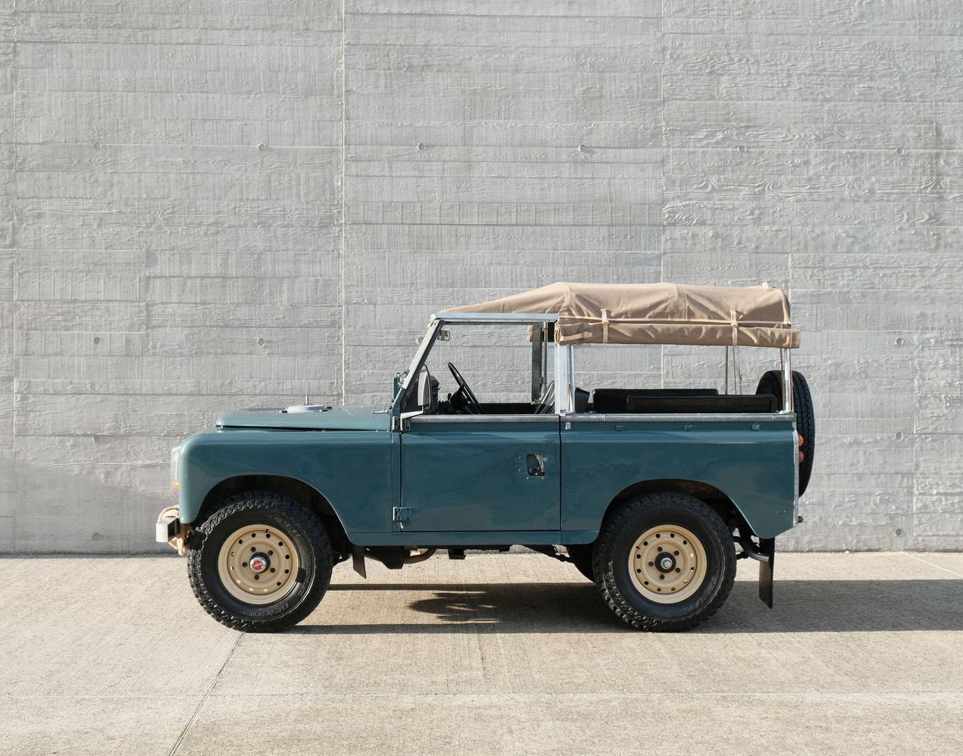 Land Rover vintage bleue avec toit et roues beiges, garée contre un mur en béton.