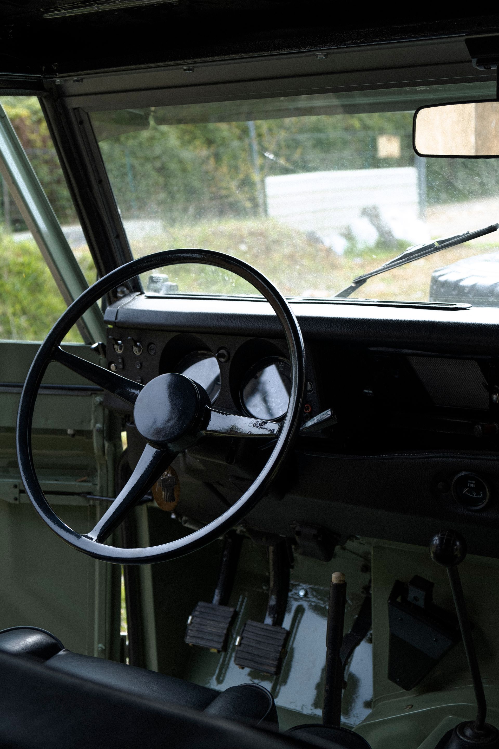 Intérieur d'un véhicule ancien : volant, tableau de bord, pédales, pare-brise et rétroviseur noirs.