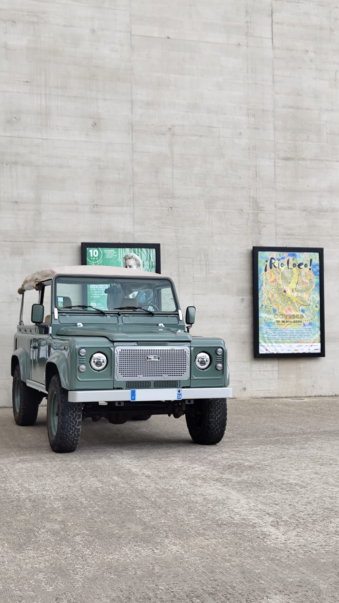 Un Land Rover Defender vert est garé à côté d'un mur en béton orné d'affiches encadrées.