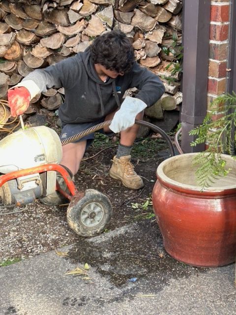 Man uses drain cleaning machine by a planter and stacked wood.
