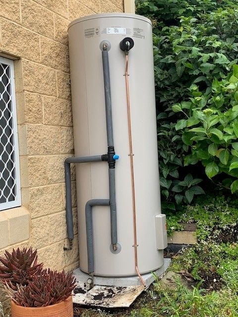 Tan water heater against a brick wall, with plumbing and a garden setting.