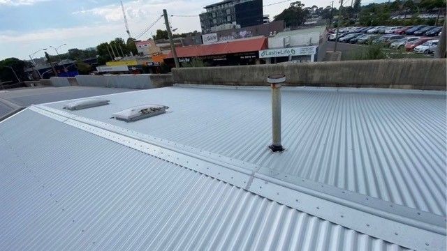 Metal corrugated roof with a vent, two skylights, and buildings in the background.