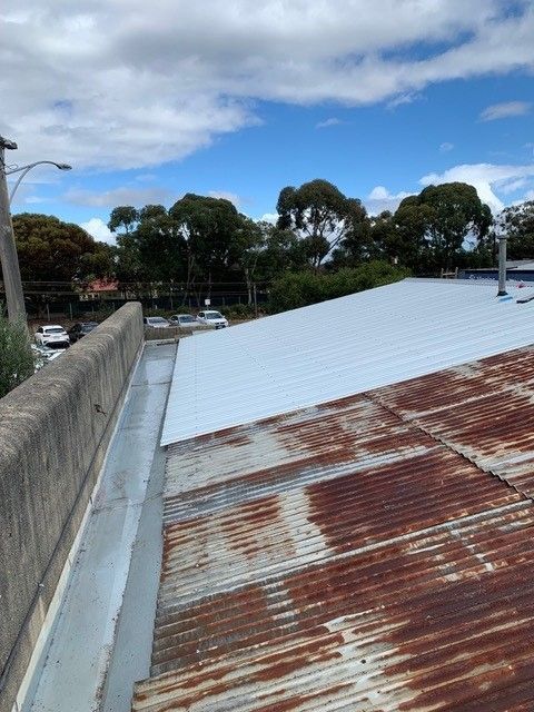 A rusty, corrugated metal roof with a newer, silver section. A concrete ledge and trees are in the background.