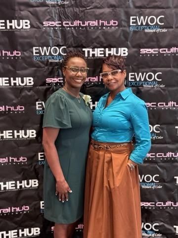 Two people smiling in front of a THE HUB and EWOC event backdrop, one in green and one in blue and brown.