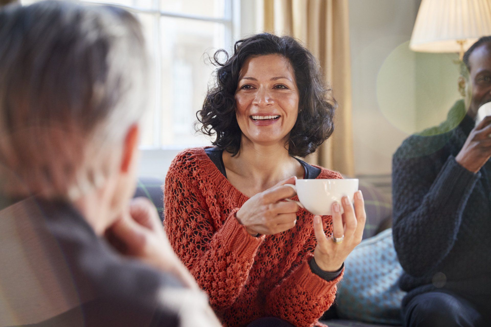 Three people sit together indoors, chatting and holding mugs in a cozy, relaxed setting.