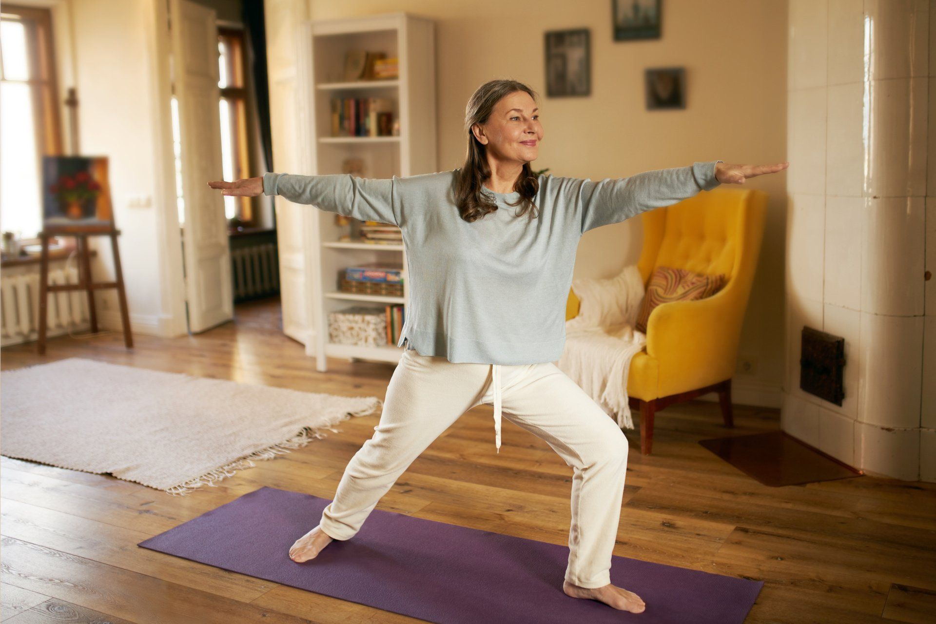 Person in a light blue long-sleeve shirt and white pants performing a Warrior II yoga pose on a purple mat in a living room.