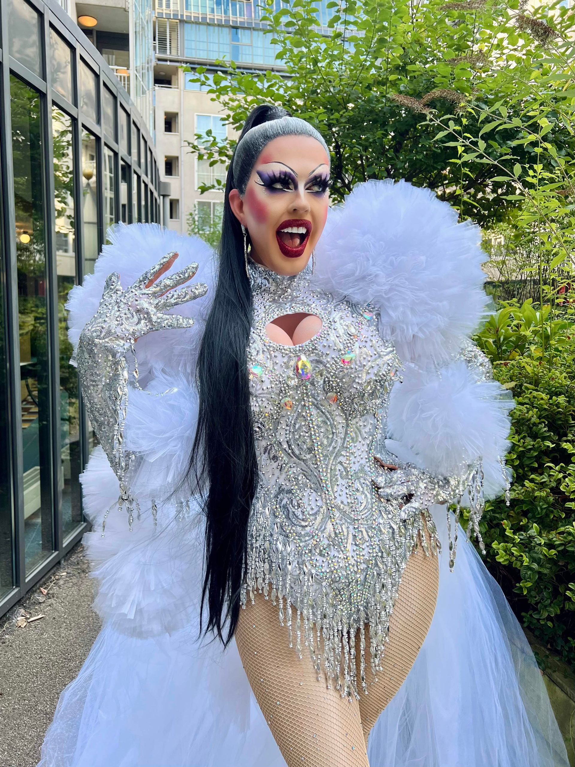 Een drag queen in een zilverkleurige outfit met pailletten, witte mouwen en sleep van veren, staat buiten bij een gebouw.