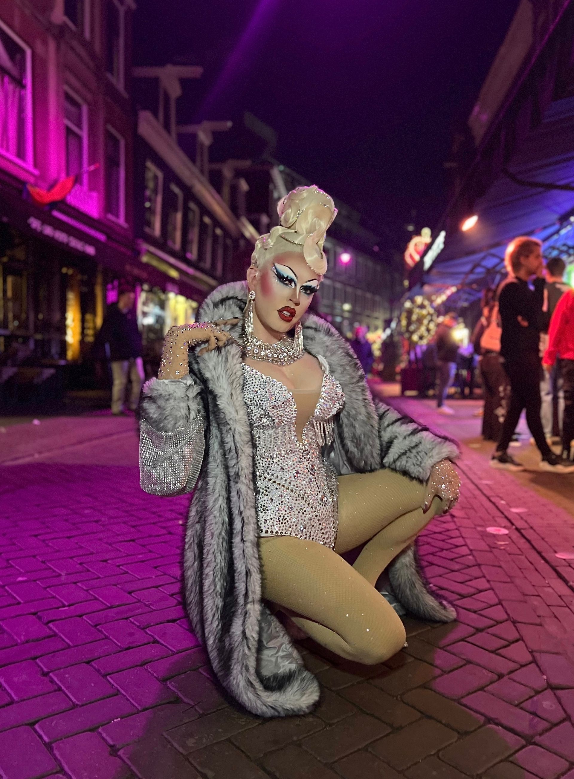 Drag queen in een zilverkleurige outfit poseert 's nachts op een neonverlichte straat.