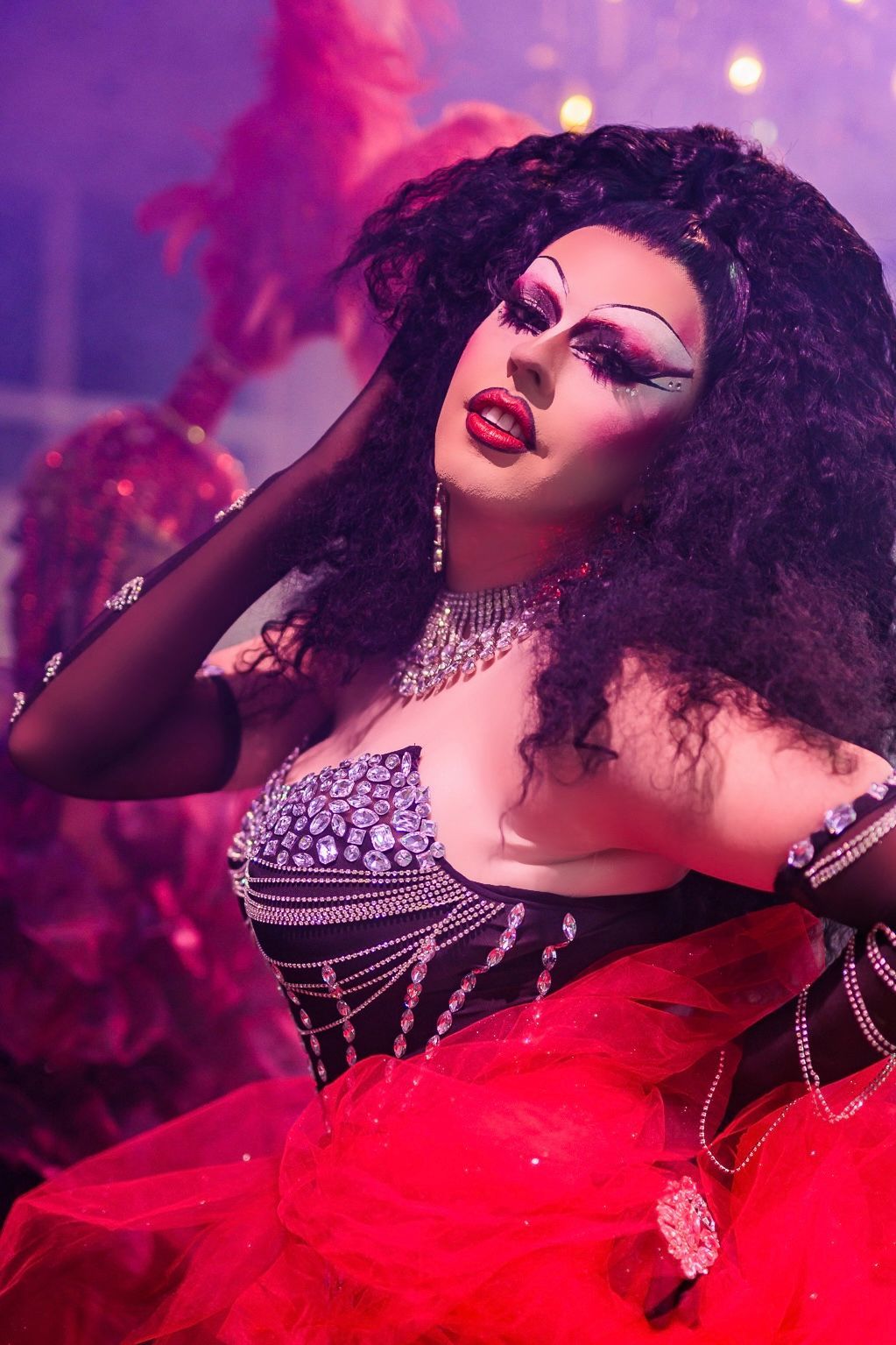 Een glamoureuze drag artiest in een rode tulejurk en een glinsterend topje poseert onder roze-paarse podiumverlichting.