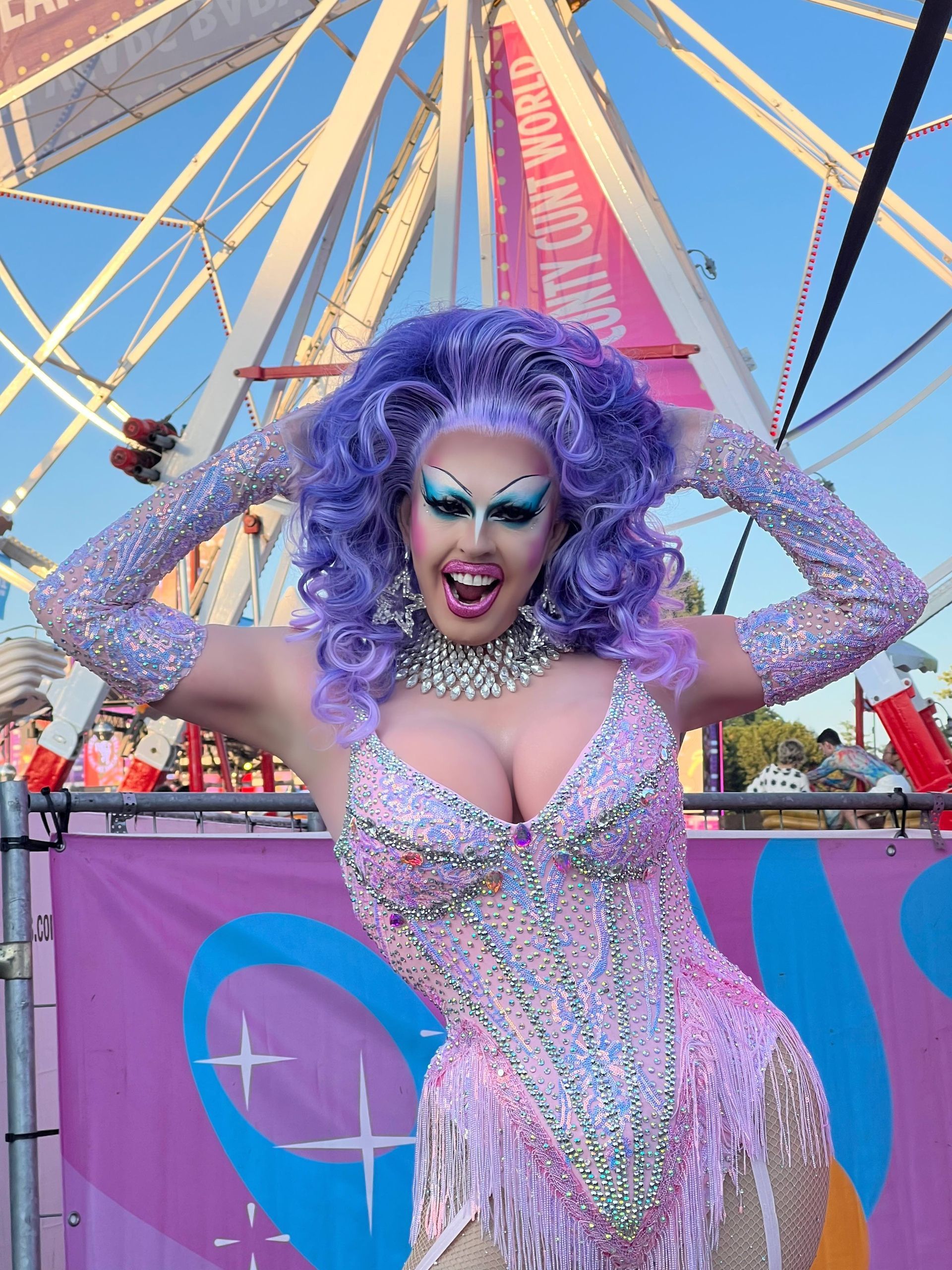Een drag artiest in een sprankelend lavendelkleurig bodysuit en pruik poseert met een reuzenrad op de achtergrond.