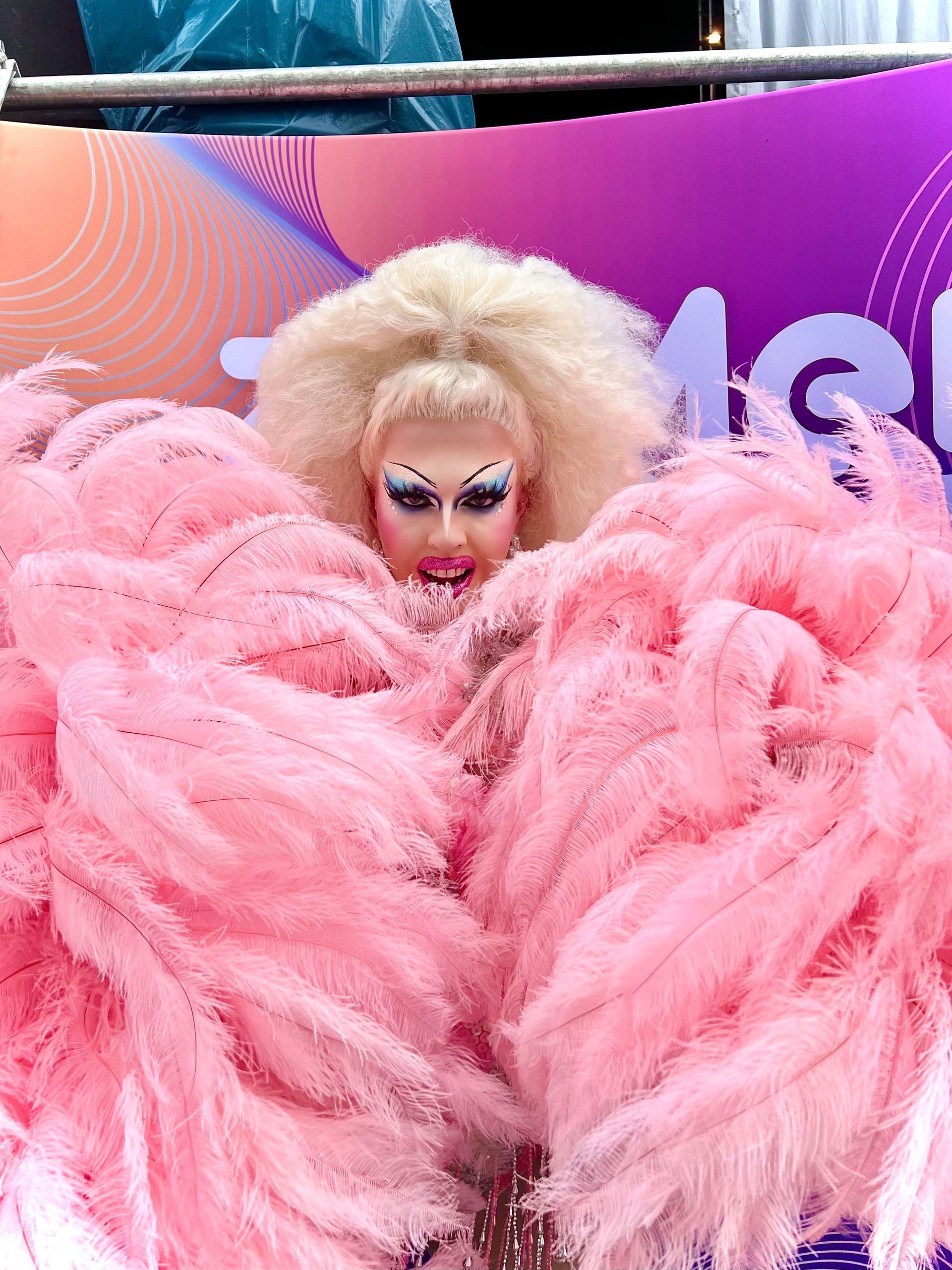 Persoon in een roze verenkostuum met blond haar en opvallende make-up op een kleurrijke podiumachtergrond.