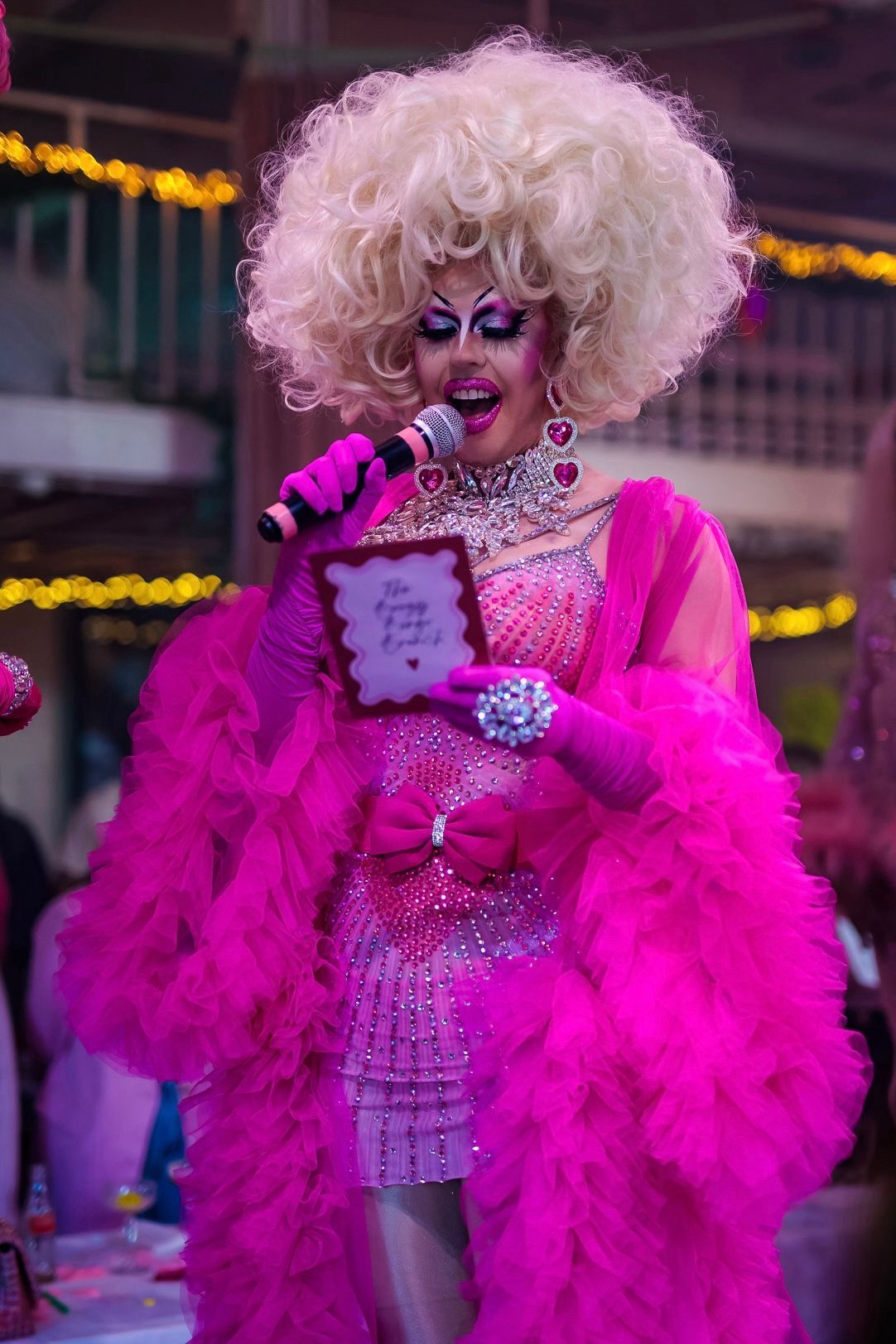 Een drag queen in een felroze outfit met veren zingt op het podium in een microfoon en houdt een klein bordje vast.