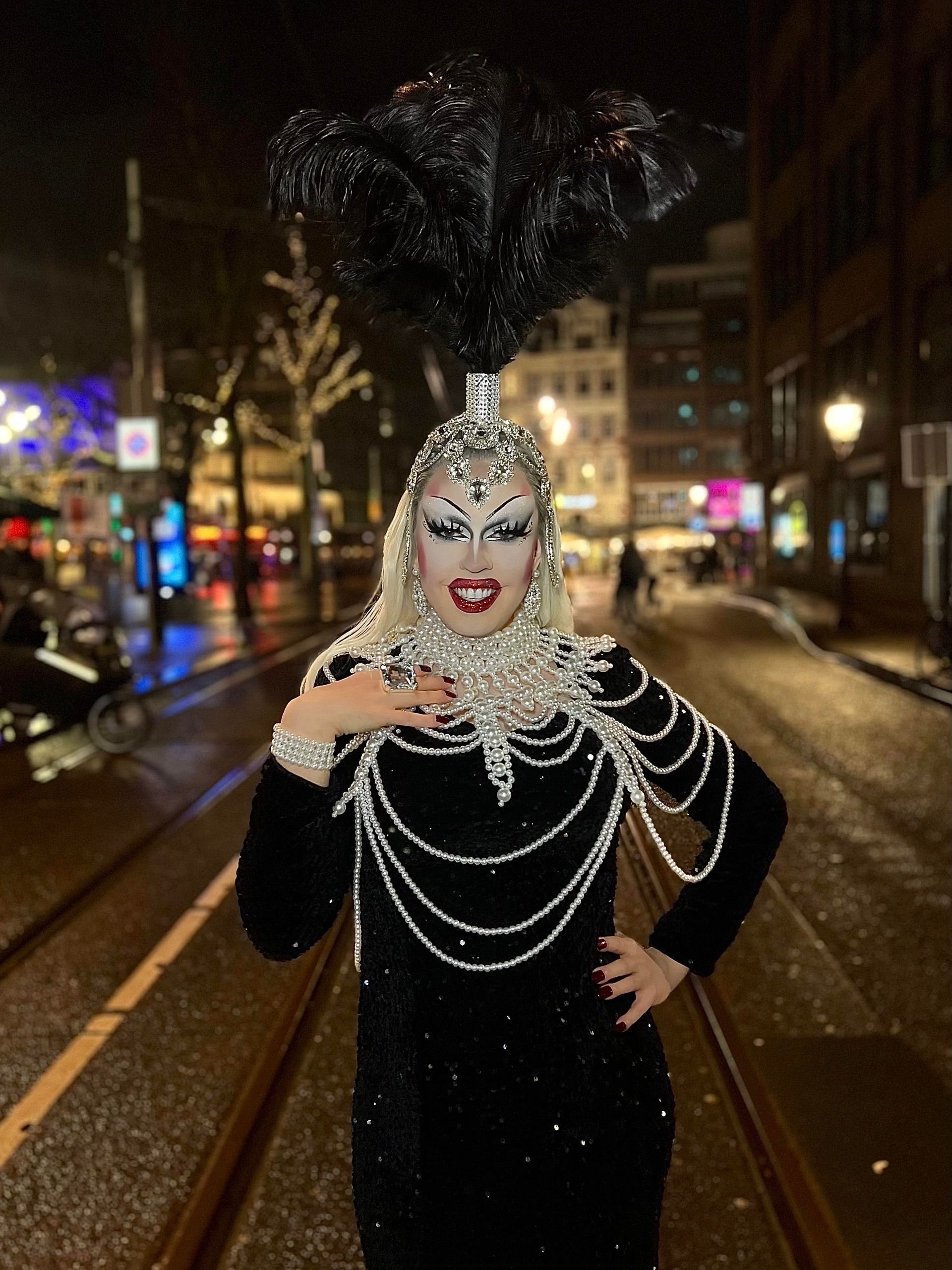 Een drag artist in een zwarte outfit met pailletten en een met hoofdtooi poseert 's nachts op een natte stadsstraat.