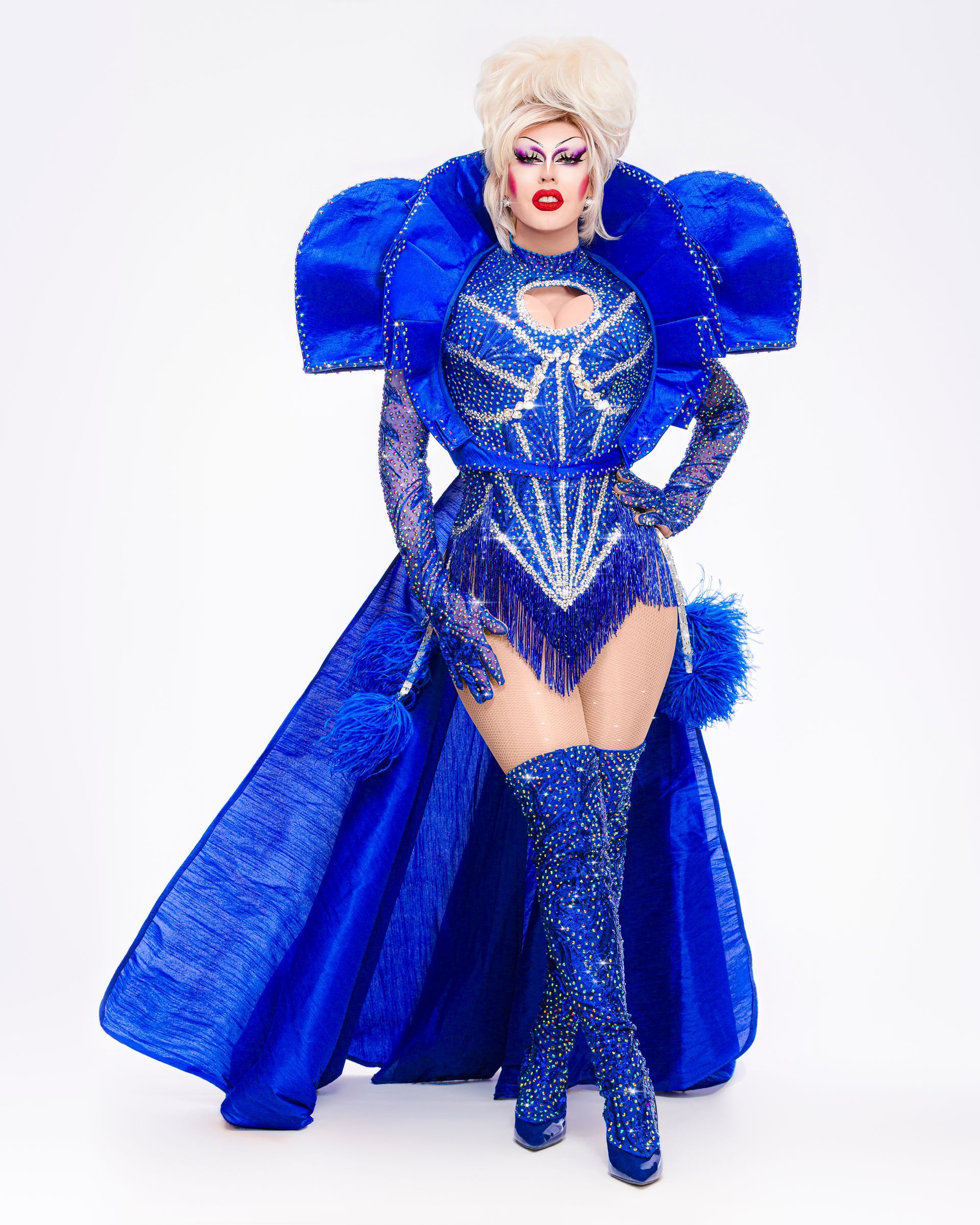 Een drag queen in een glinsterend blauw kostuum met oversized schouders en laarzen, poserend tegen een witte achtergrond.