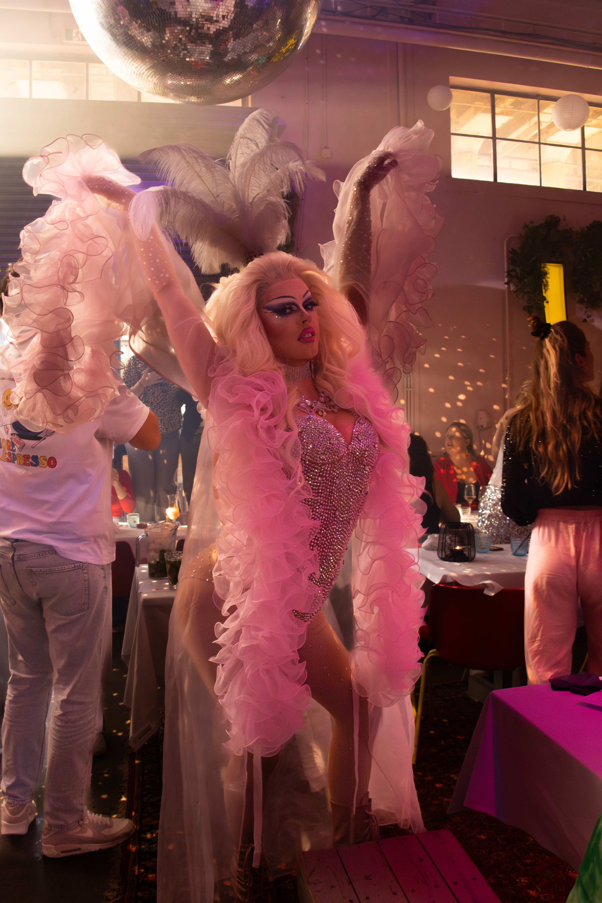 Een drag artiest met roze verenboa en zonnebril poseert op een glamoureus feest onder een discobal.