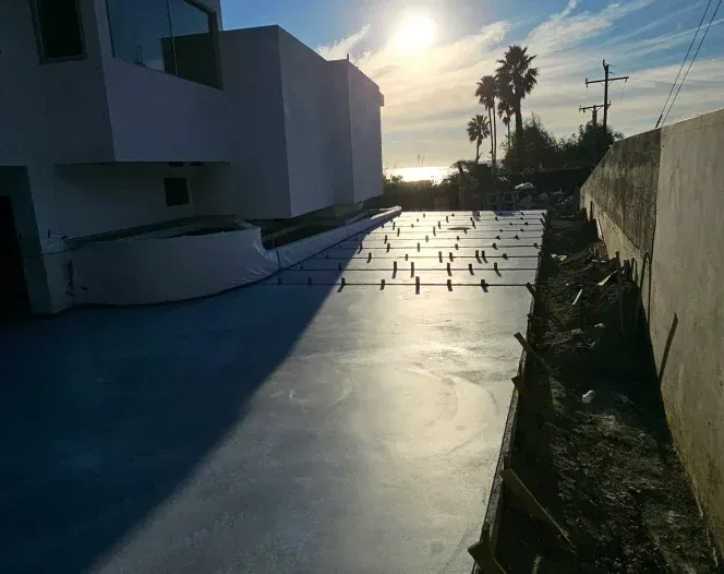 Sunlit rooftop with fresh concrete slab and rebar, beside a white building and fence