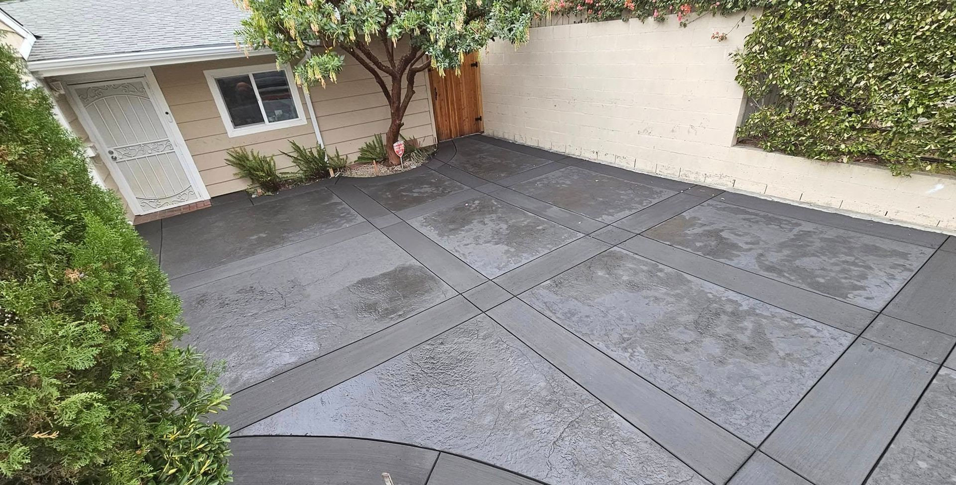 Wet gray stone patio with geometric pavers beside a house and hedge-lined wall