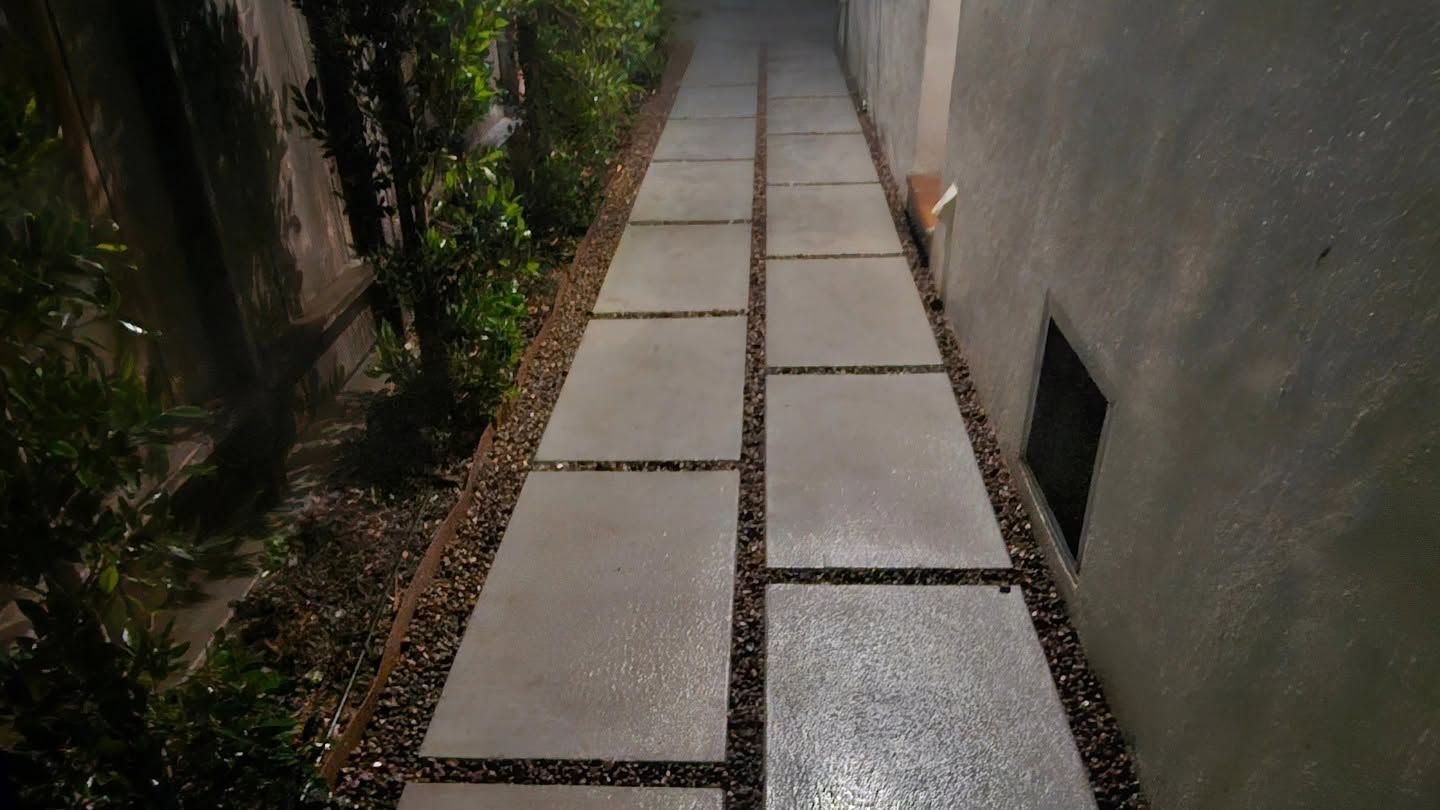 Narrow concrete walkway between walls, with small garden on the left and a drain opening on the right.