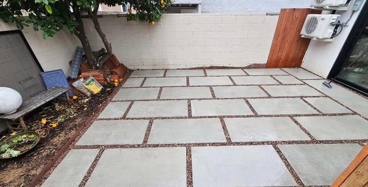 Backyard patio with large rectangular stone pavers, bordered by plants and a wooden gate.