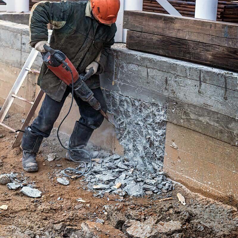 Worker using a jackhammer to break concrete beside a wall, with debris scattering on the ground.
