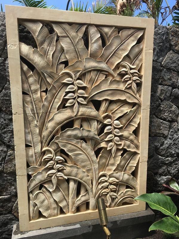 Stone relief panel with tropical leaves and flowers, framed in beige stone outdoors by rocks and plants