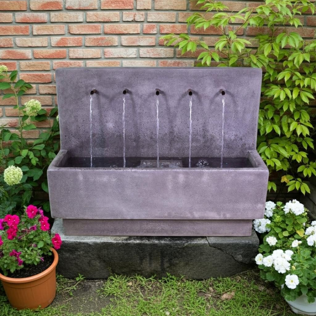 Gray garden fountain with five water streams, set against a brick wall and surrounded by flowers and greenery