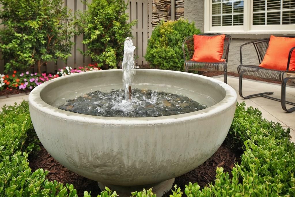 Outdoor stone fountain with water spraying into a landscaped garden beside a patio bench with orange cushions