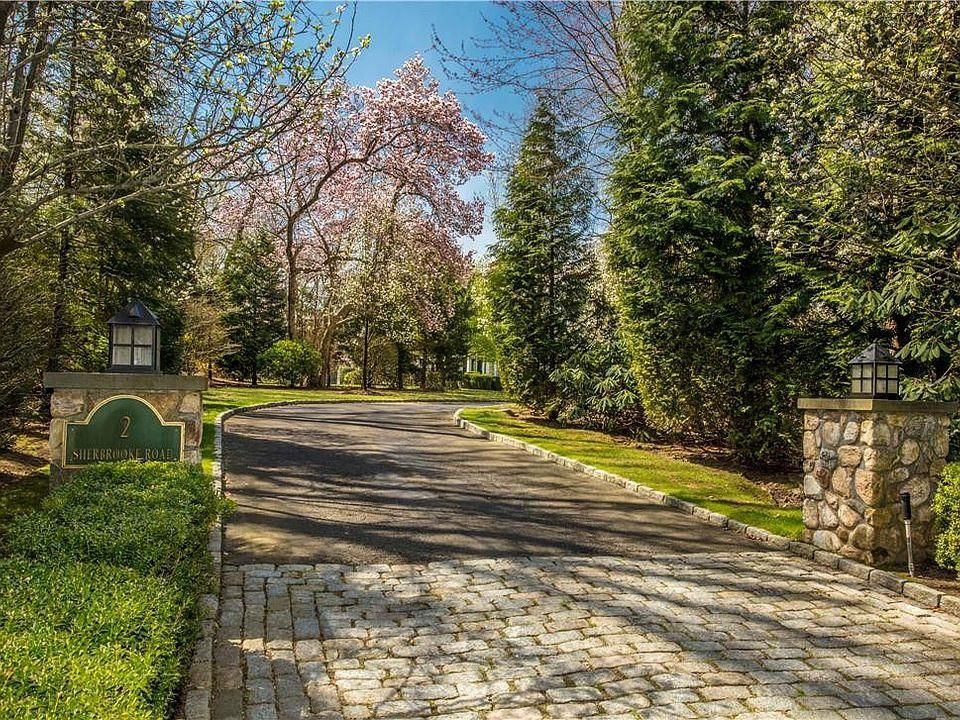Stone driveway through a landscaped garden with tall evergreen trees and pink blossoms in spring.