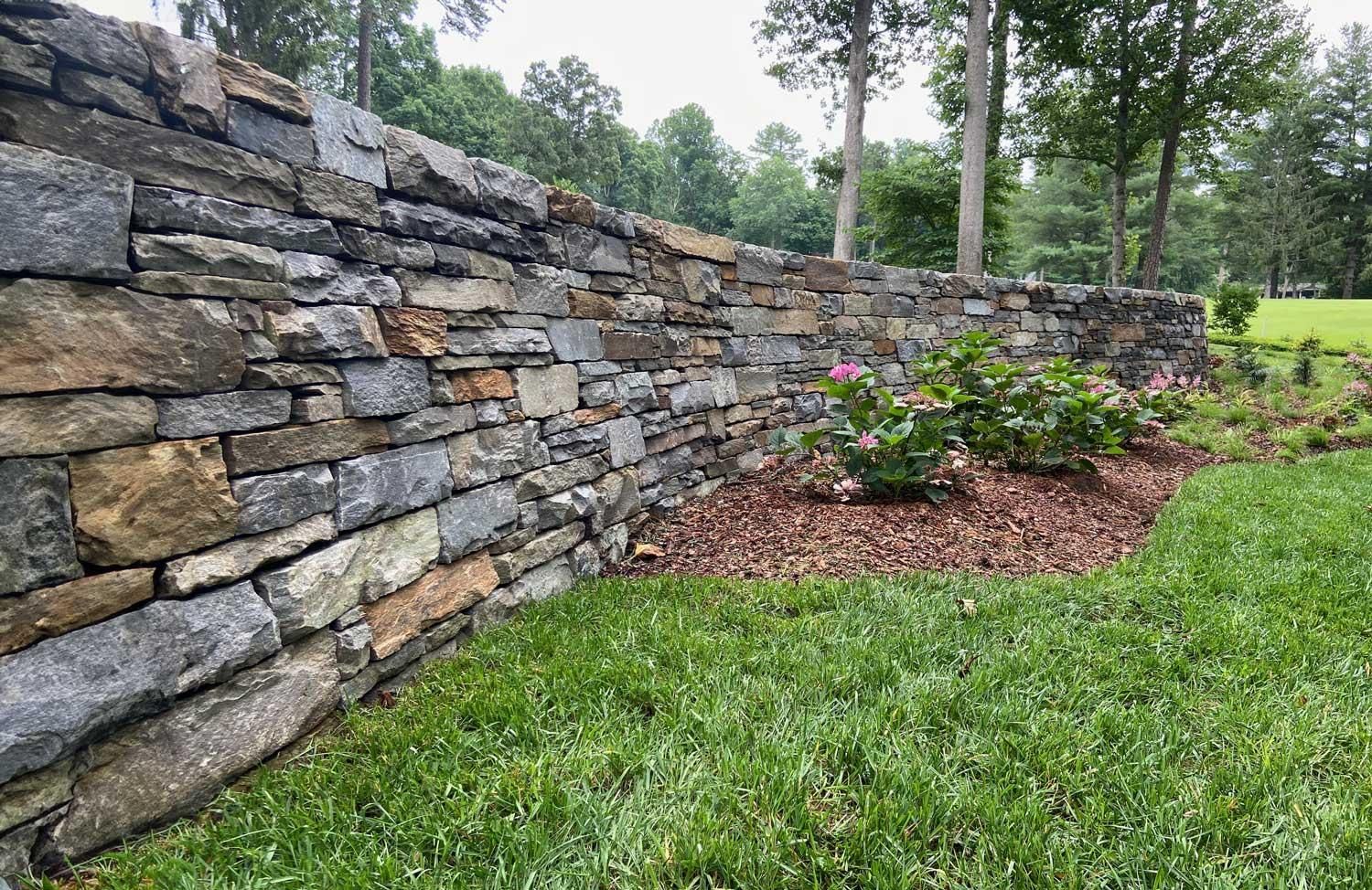 Stone retaining wall beside green lawn and mulched flower bed with pink blooms in a wooded yard