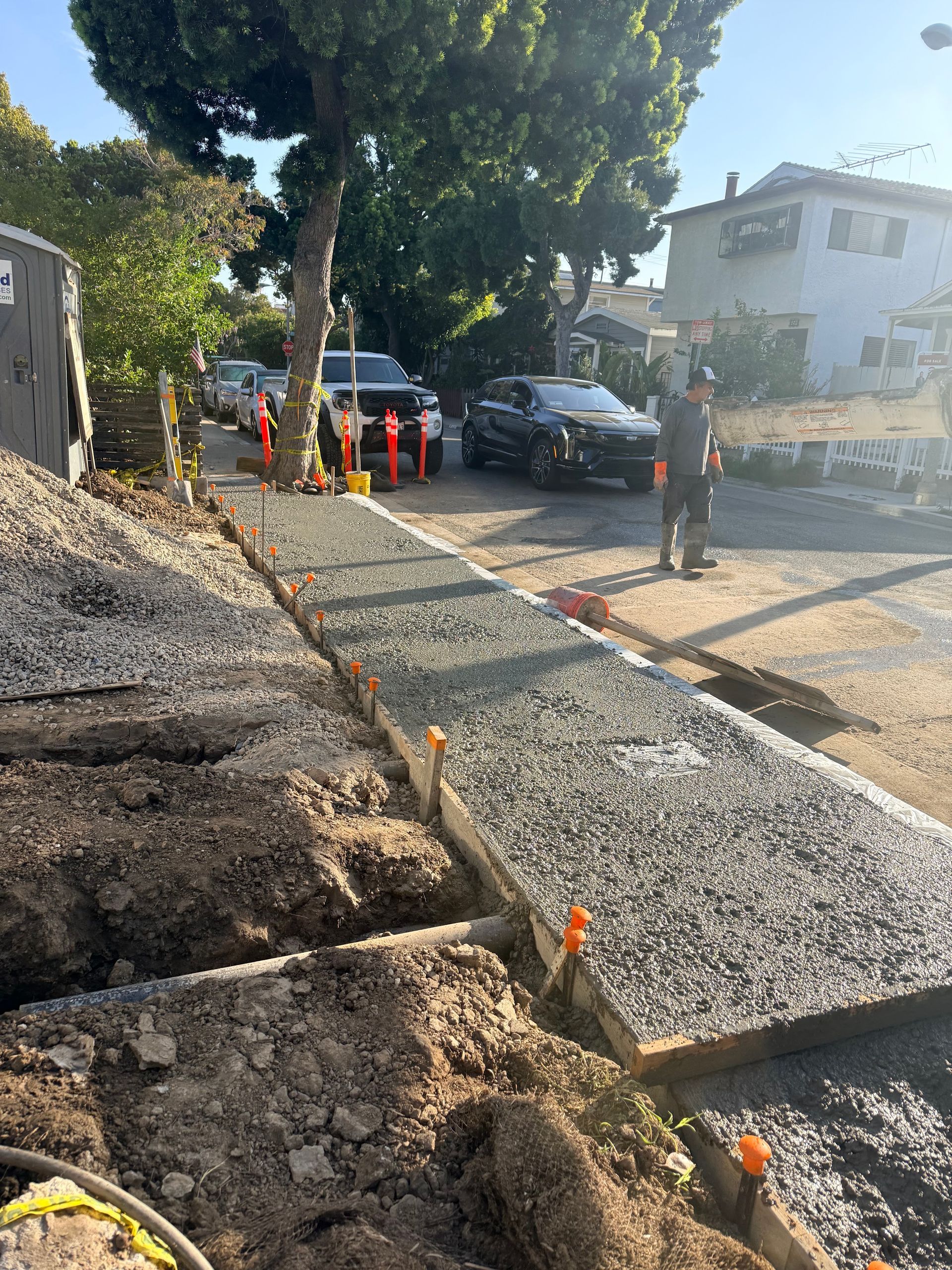 Roadside utility trench with gravel, orange cones, and workers near parked cars under trees