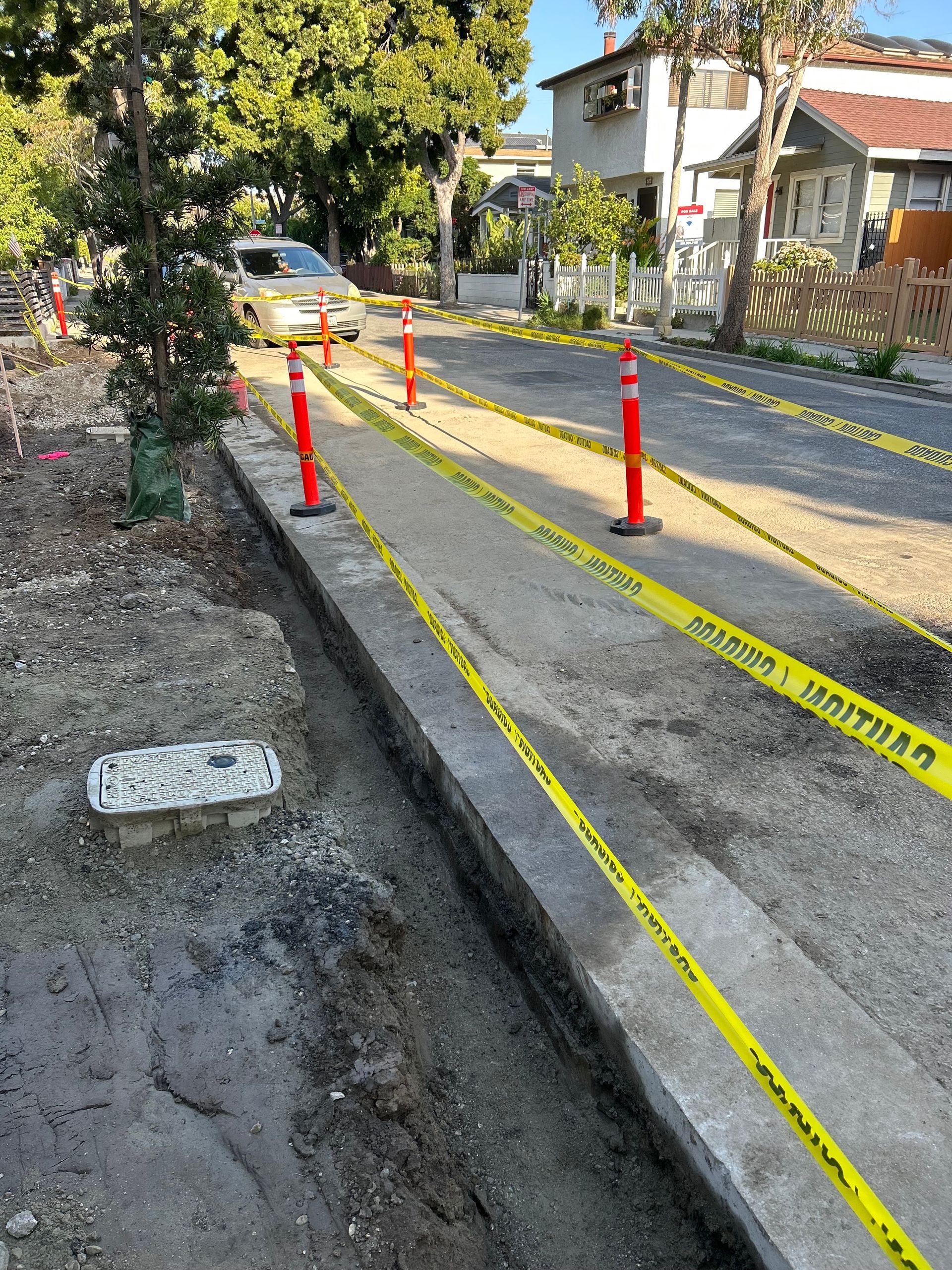 Sidewalk construction area with yellow caution tape and orange cones along a gravel path.