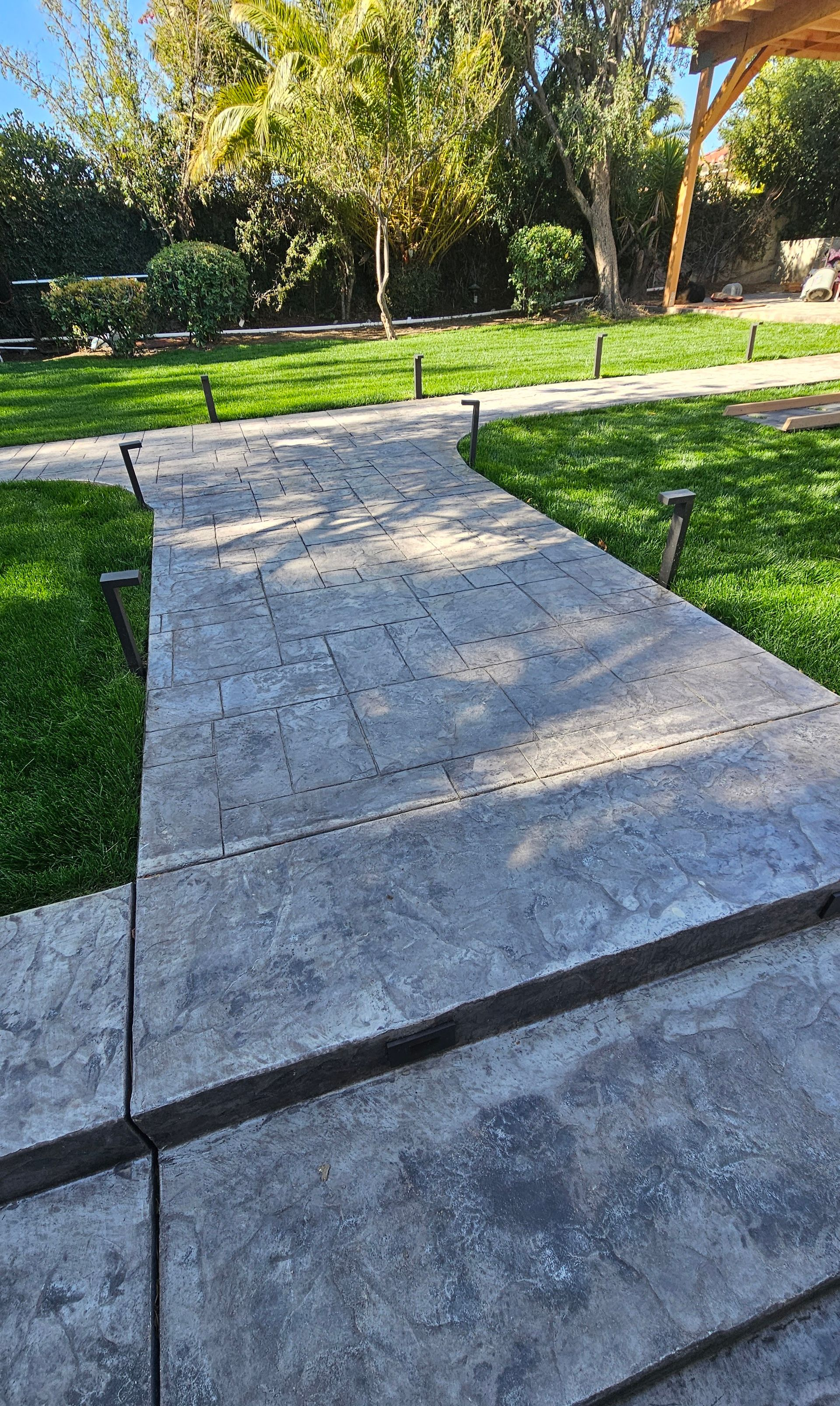 Concrete walkway leading through a sunlit park with grass, trees, and a road in the background