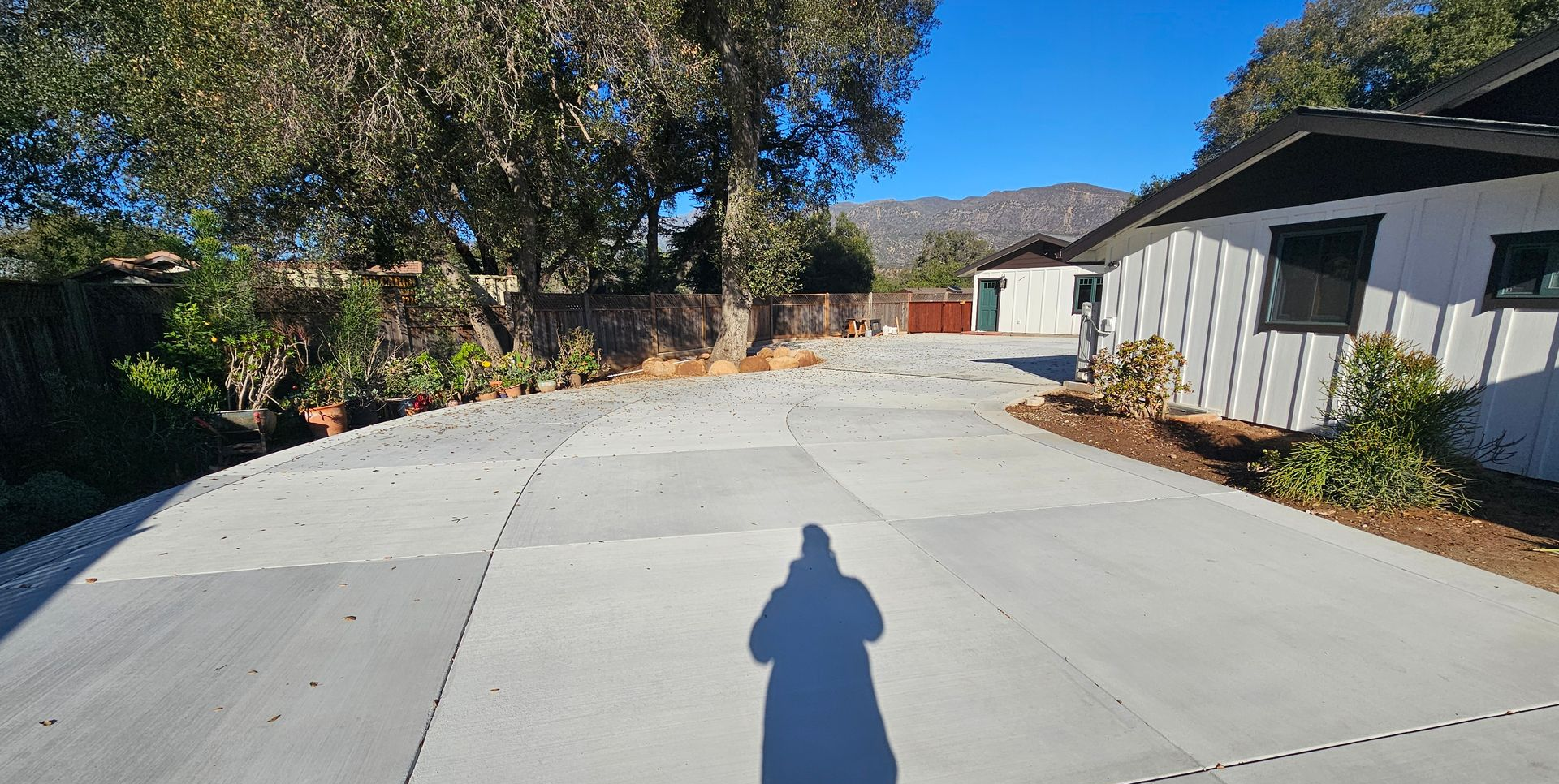 Wide concrete driveway beside houses and trees, with a person’s shadow in the foreground.