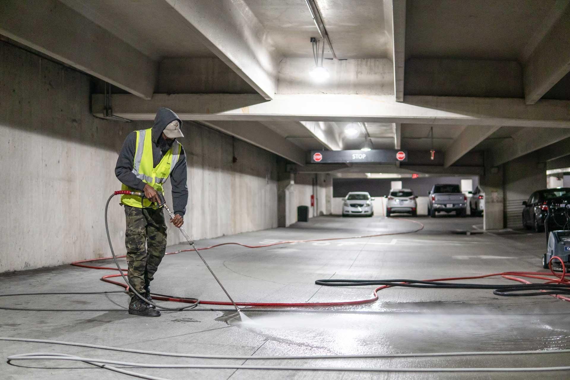 Worker pressure-washing a concrete floor in a parking garage, wearing a safety vest and helmet
