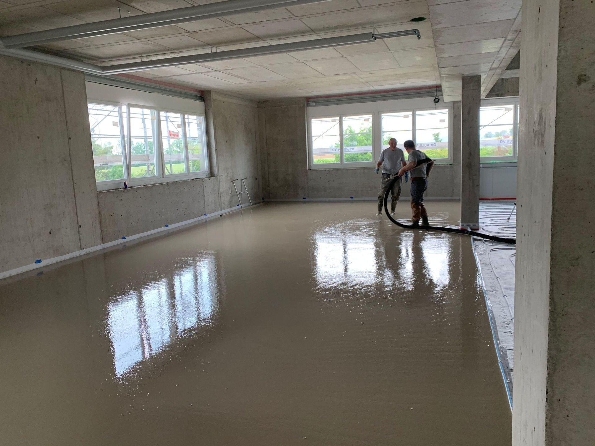 Workers smoothing wet concrete floor in a bright, unfinished room with large windows.