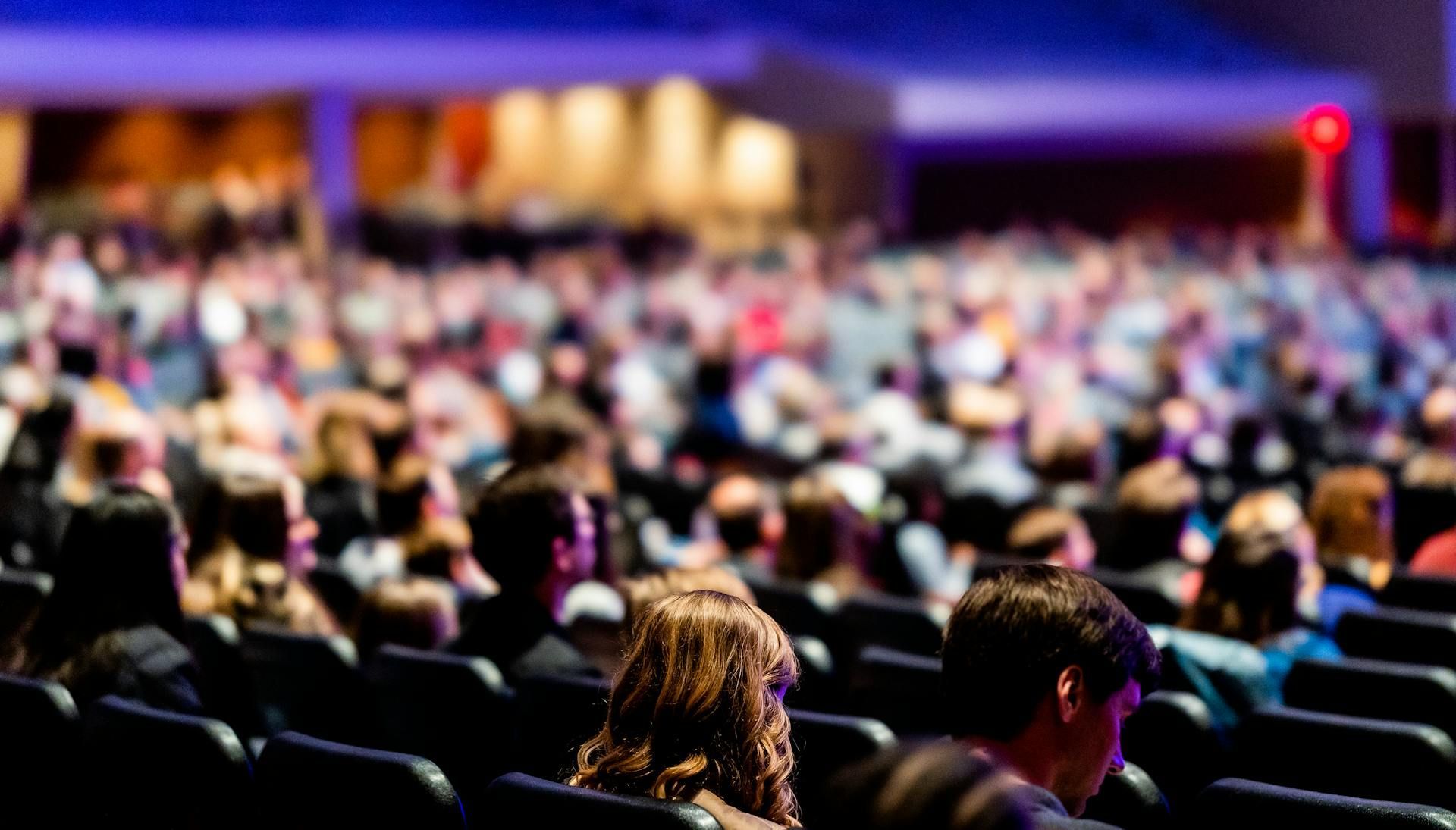 Audience seated in a large auditorium, watching a performance under blue and purple stage lights.