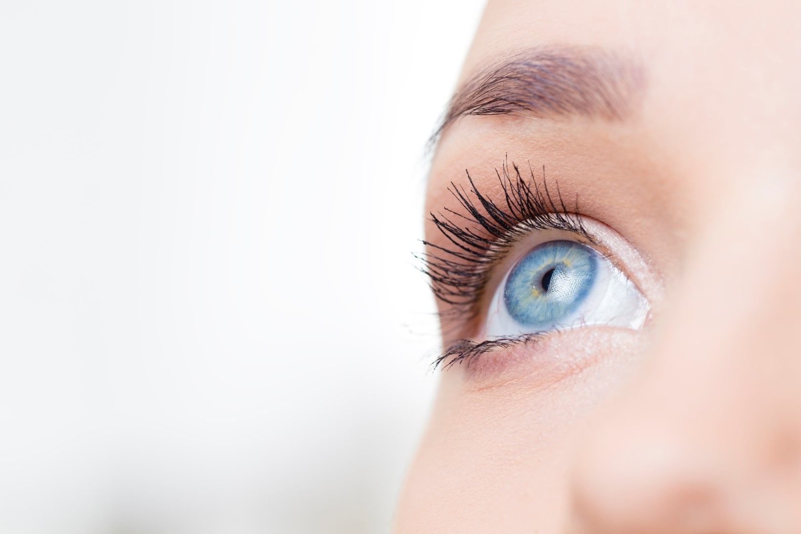 Close-up of a person's light blue eye looking upwards, with dark mascara and a light eyebrow against a white background.