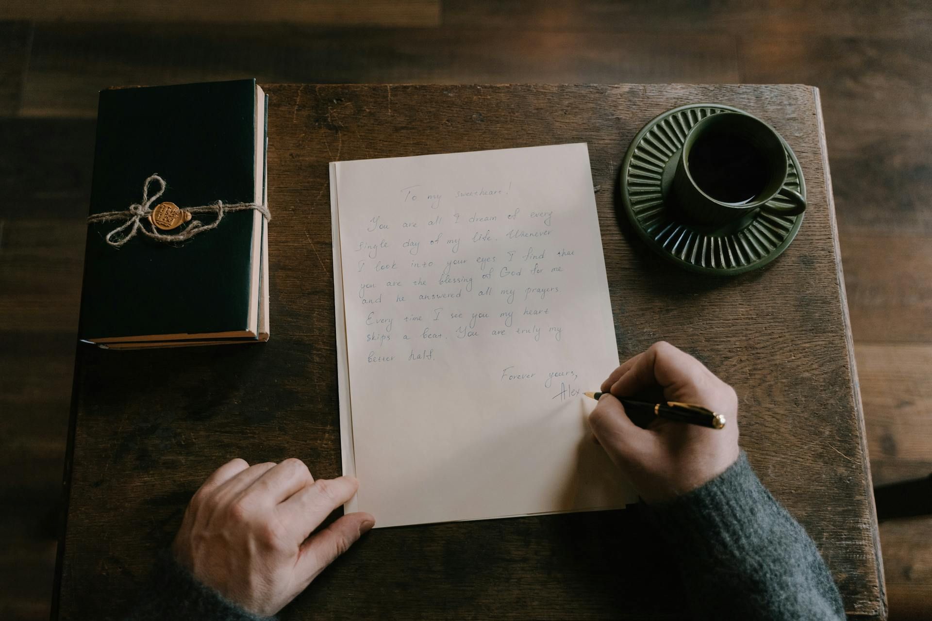 A person's hands writing on paper at a wooden desk, beside a book and teacup.