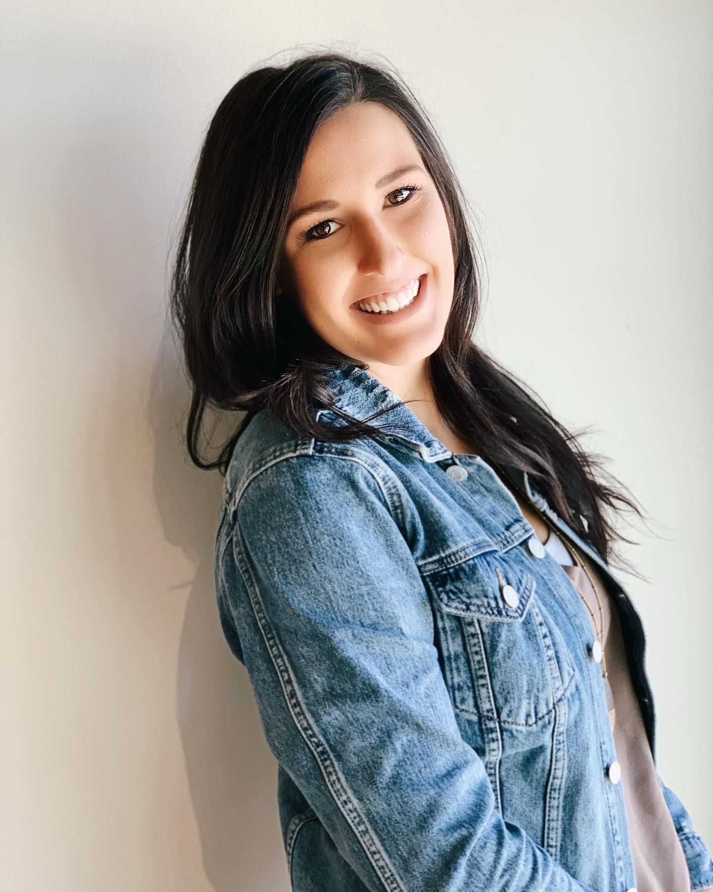A woman in a denim jacket is smiling and standing in front of a white wall.