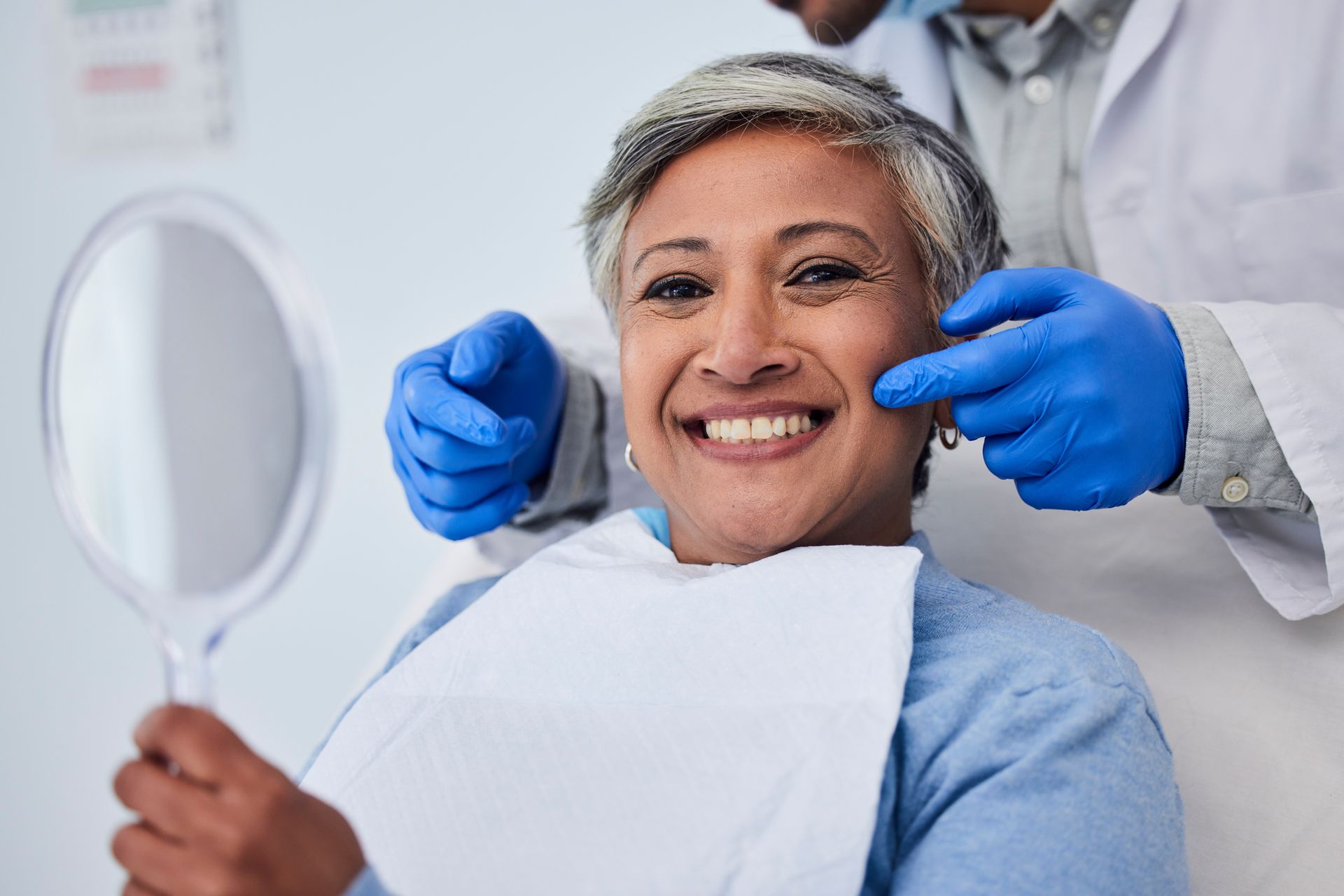 Smiling patient in dental chair with mirror while dentist adjusts blue-gloved hands nearby Smiling patient in dental chair with mirror while dentist adjusts blue-gloved hands nearby