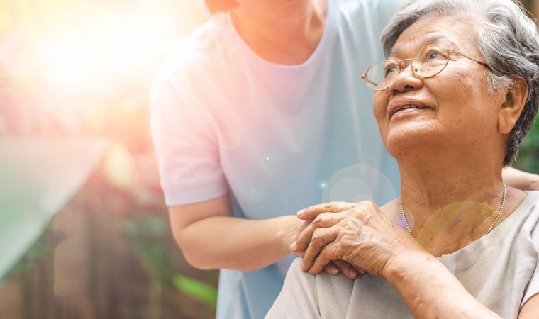 Elderly woman with glasses smiles, hand on shoulder, looking up at someone. Another person is behind her.