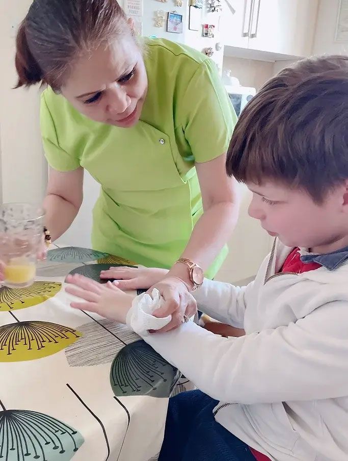 Woman in green shirt tends to boy's hand at a table, holding a wet wipe.