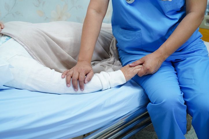 Nurse in blue scrubs holding patient's hand on a hospital bed.