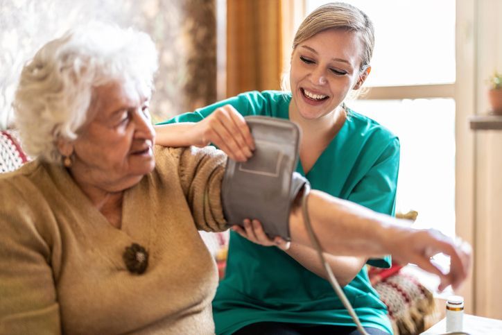 Caregiver taking an elderly person's blood pressure. Both are smiling inside a home.