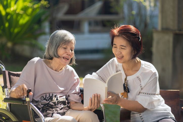 Woman in wheelchair and another woman read a book outdoors, both smiling.