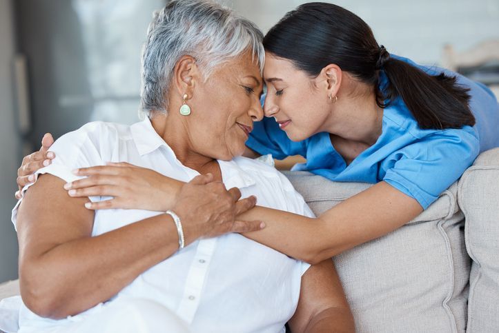 Woman hugs another woman, touching foreheads. Both are indoors, on a couch.